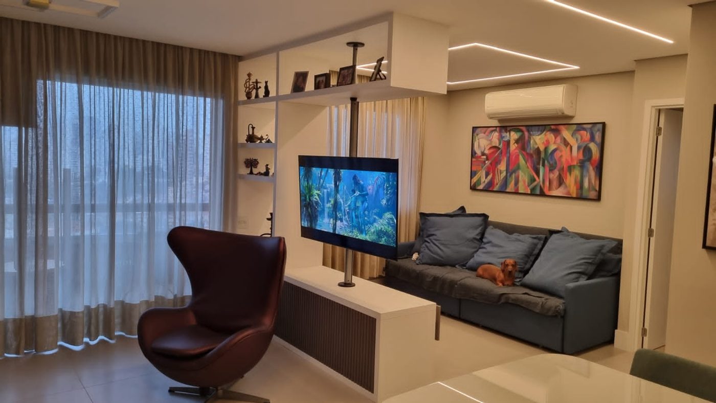 Sala com poltrona de couro, sofá cinza, TV em painel giratório e estante iluminada