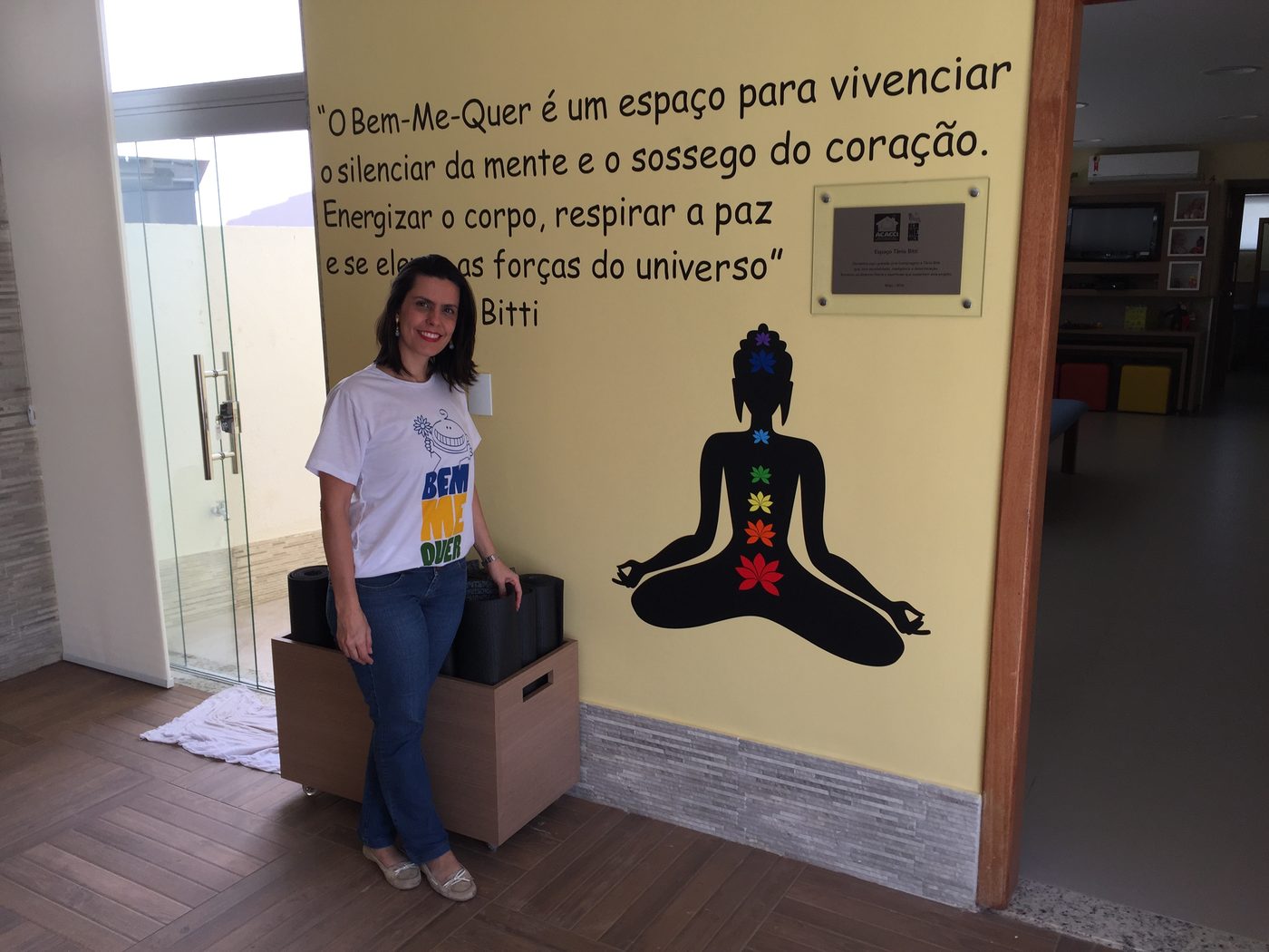 Simone Afonso ao lado de uma parede amarela com frase sobre o Bem Me Quer e silhueta de meditação com chacras coloridos